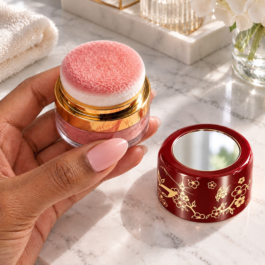 Hand holding an open blush jar with its puff applicator, the red floral lid with its mirror on the counter
